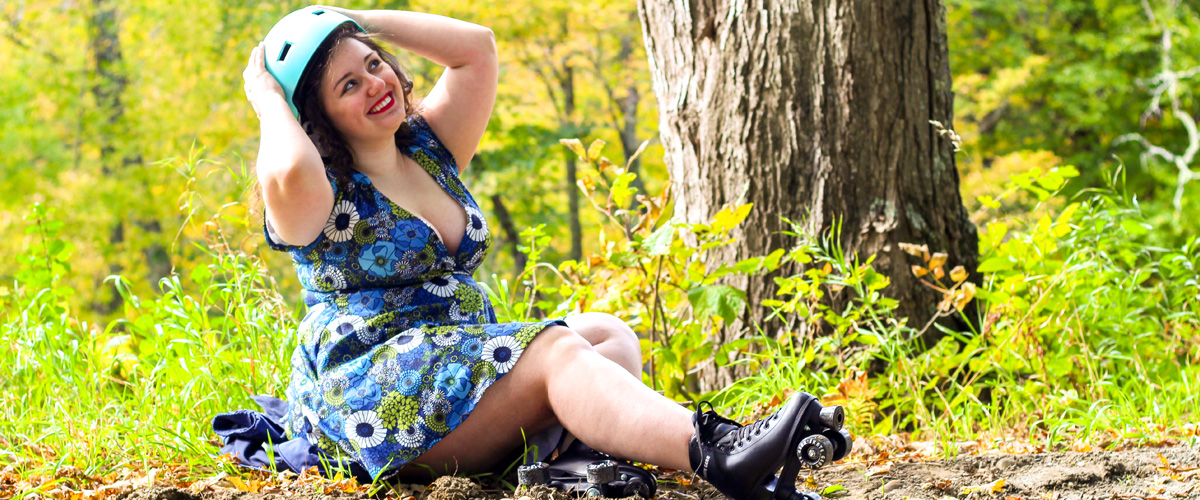 A photo of Joanna No Banana, who is the writer of this blog post, a white woman with brown, curly hair wearing a blue floral dress and black roller skates while putting on a helmet